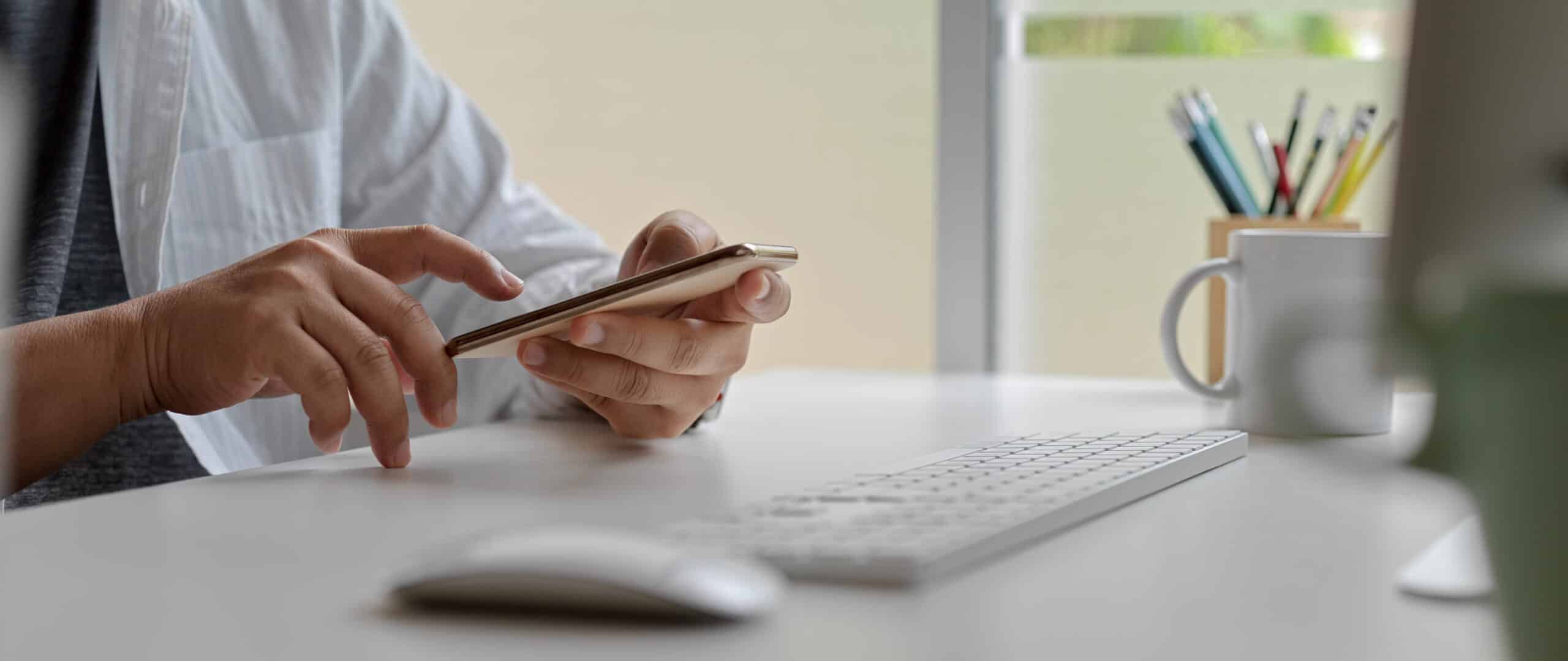 Male worker using smartphone while sitting at office desk with computer device and other supplies
