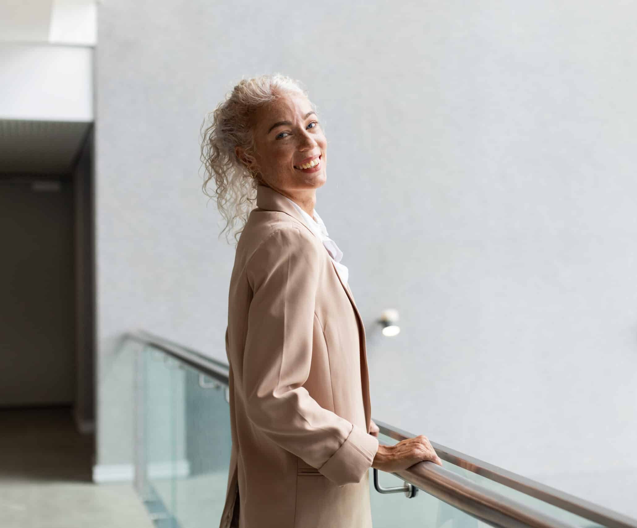 Smiling woman in business attire standing by railing in modern office building, copy space. Businesswoman, professional, corporate, architecture, confident